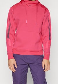 Rozpinana bluza z kapturem w jasnoróżowym kolorze, o teksturowanym korpusie, gładkich rękawach, przedniej kieszeni i regulowanymi sznurkami. Połączona z fioletowymi spodniami.