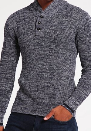 Mann trägt einen grauen, strukturierten Strickpullover mit geknöpftem Kragen und dunkelblaue Jeans, Hand ruht in der Tasche.