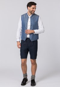 Blau gemusterte Weste mit hohem Kragen über einem weißen Hemd; kombiniert mit dunklen Shorts mit bestickten Details und silbernen Knopfakzenten.