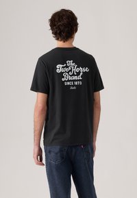 Černé bavlněné tričko s velkým bílým potiskem na zádech se slovy "The Two Horse Brand" a "Since 1873." Krátké rukávy, volný střih.