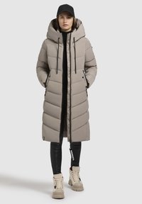 Hellgraue Pufferjacke mit Kapuze, ausgestattet mit einem Frontreißverschluss, Seitentaschen und horizontalen Nähten. Getragen über schwarzen Leggings und beige Stiefeln.