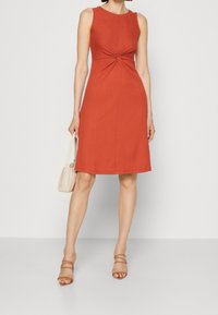 Robe sans manches couleur rouille avec un détail noué à la taille, corsage ajusté, jupe évasée et texture douce. Accompagnée d'un petit sac à main beige.