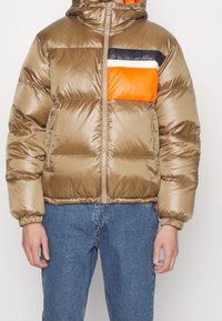 Homme portant une doudoune brillante marron avec des détails color block noir, blanc et orange, associée à un jean bleu.