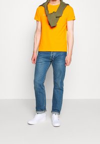 Camiseta de algodón naranja con cuello redondo, combinada con jeans de mezclilla azules. Un suéter verde está drapeado sobre los hombros. Zapatillas blancas completan el look.