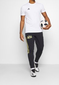 Vit kortärmad sporttröja med svart logotyp, kombinerad med mörka joggers med gula ränder och grafisk design, hållande en fotboll.