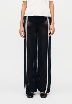 Čierne sieťované nohavice s voľným, širokým strihom, s bielymi bočnými pásmi a textúrovaným vzorom. Nosené s priliehavým bielym topom.