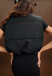 Schwarze, wattierte Weste mit rundem, voluminösem Design, ausgestattet mit einer glatten Textur und elastischem Bund, getragen über einem enganliegenden schwarzen Oberteil.