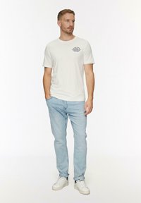 Weißes Baumwoll-T-Shirt mit kurzen Ärmeln, versehen mit einem navy-blauen Grafikdesign auf der Brust. Kombiniert mit hellblauen Jeans und weißen Sneakers.