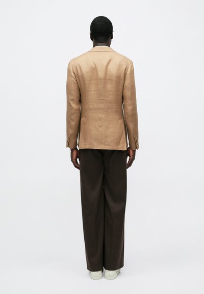 Άνδρας που στέκεται με την πλάτη γυρισμένη, φοράει μπεζ σακάκι, σκούρα καφέ παντελόνια με φαρδιά πόδια και λευκά αθλητικά παπούτσια σε απλό λευκό φόντο.