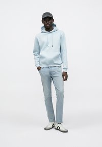 Lys blå hettegenser med frontlomme, kombinert med lys blå jeans og svart-hvite joggesko. Svart cap med preget logodetalj.