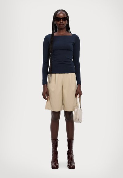 Marine langermet topp, beige plisserte shorts og brune knelang støvler. Tilbehør inkluderer en liten lys bag og mørke solbriller.