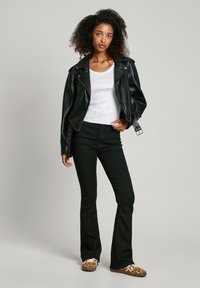 Black leather biker jacket, white t-shirt, black flared pants, and leopard-print shoes. Curly hair, hands on hips, neutral background.