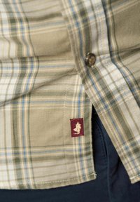 Bruine geruite shirt met groene en blauwe strepen, knoopdetails en een bordeauxrood logo-op-natuur aan de zoom. De stof heeft een gestructureerde uitstraling.