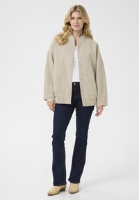 Veste beige surdimensionnée avec un col rond, boutons frontaux visibles, deux grandes poches et ourlet côtelé, associée à un jean skinny sombre et des bottes beiges.