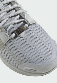 Szare buty sportowe z siateczkowym wierzchem, wyraźnymi otworami wentylacyjnymi, eleganckim designem i metalowymi akcentami. Posiadają sznurówki i detal z logo.