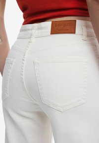 Weiße Denim-Jeans mit figurnahem Schnitt, ausgestattet mit einem braunen Lederpatch mit der Aufschrift "Desigual" und zwei Gesäßtaschen mit sichtbaren Nähten.