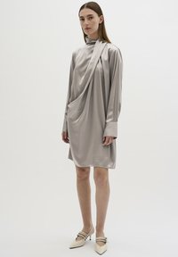 Zijdegrijze jurk met lange mouwen, gedrapeerd stofontwerp, hoge kraag en een losse pasvorm. Vergezeld van puntige beige hakken met bandjes.
