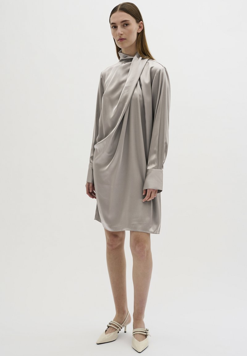 Zijdegrijze jurk met lange mouwen, gedrapeerd stofontwerp, hoge kraag en een losse pasvorm. Vergezeld van puntige beige hakken met bandjes.