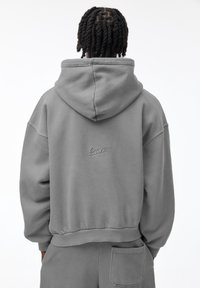 Sweat à capuche gris avec cordon de serrage, coupe ample, poignets et ourlet côtelés. Présente un petit logo au dos. Fabriqué en coton doux.