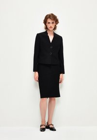 Czarna dopasowana marynarka z dekoltem w kształcie litery V, z dwoma złotymi guzikami, w połączeniu z czarną ołówkową spódnicą do kolan. Gładki materiał o ustrukturyzowanym kroju.