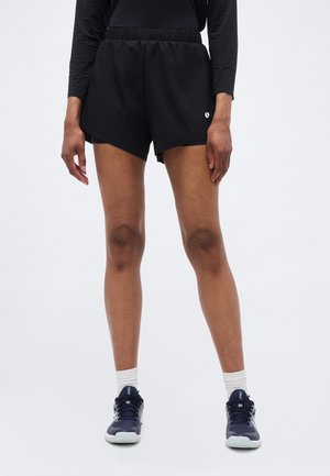 Fekete sportnadrágot, fekete hosszú ujjú felsőt, fehér zoknit és tengerészkék futócipőt viselő személy, egyszínű háttér előtt állva.