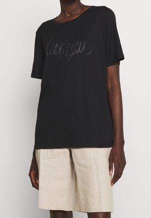 Personne portant un T-shirt noir à manches courtes avec un texte pailleté subtil et un short beige arrivant aux genoux, debout devant un fond blanc uni.