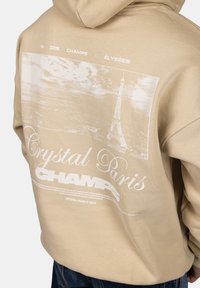Béžová mikina s kapucí, která na zadní straně nese bílou grafiku Eiffelovy věže a text "Crystal Paris CHAMP", s jemnou a hladkou strukturou.