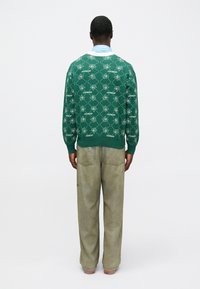 Suéter verde de malha com um padrão floral e texto, combinado com jeans de denim de corte solto e de cor clara. Design minimalista.