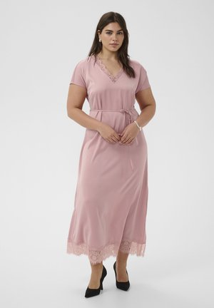 Femme portant une robe midi rose ornée de dentelle, à manches courtes, avec une ceinture à la taille, et des escarpins noirs pointus, debout devant un fond uni.