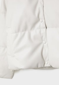 Section de veste matelassée blanche avec boutons-pression et détail de couture visible sur tissu matelassé doux.