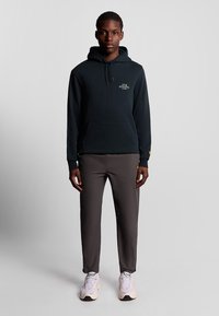 Felpa nera con tasca frontale e cordino, pantaloni grigi con dettagli gialli. Il modello indossa sneakers chiare. Abbigliamento sportivo casual.