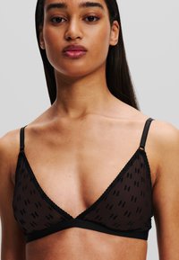 Zwarte bralette van doorschijnende stof met een herhalend logo-patroon, driehoekige cups, kantafwerking en dunne verstelbare schouderbandjes.