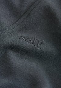 Tmavá látka s jemnou texturou, na které je vyšitý nápis 'self.' v o něco světlejší barvě. Hladký a minimalistický design.