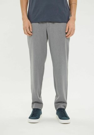 Mann iført grå rette bukser og mørkeblå joggesko står mot en hvit bakgrunn.