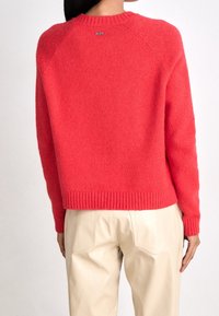 Czerwony wełniany sweter z okrągłym dekoltem, ściągaczami na mankietach i u dołu. Z tyłu znajduje się małe metaliczne logo. W połączeniu z beżowymi spodniami.
