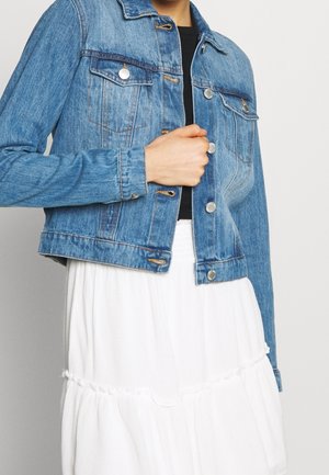 Osoba nosící modrou džínovou bundu zapnutou přes černý top a bílou vrstvenou sukni, držící bundu jednou rukou u hrudi.