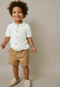 Vit texturerad kortärmad skjorta med knappar, kombinerad med bruna shorts och ljusgrå slip-on skor. Stående pose med neutral bakgrund.