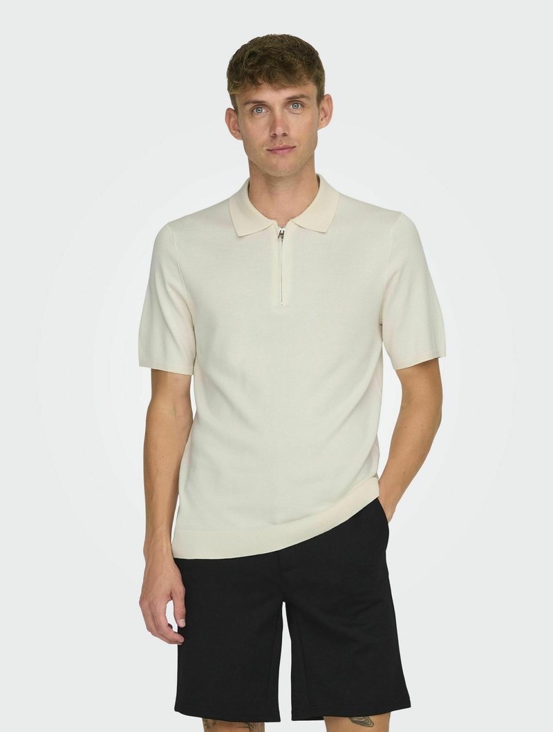 Polo de punto en beige claro con mangas cortas y cuello con cremallera, combinado con pantalones cortos negros. Textura suave, diseño ajustado.