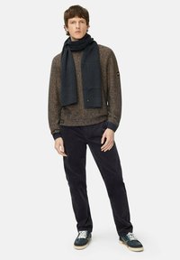 Gebreide trui in bruin en zwart tinten met geribbelde manchetten, gecombineerd met donkere broek en een donkere sjaal. Sneakers met witte veters.