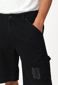 Shorts de mezclilla negros con corte recto, que presentan dobladillos deshilachados, un detalle de parche y bolsillos frontales. La tela tiene una textura suave.