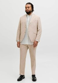 Homme aux cheveux tressés portant un costume beige, une chemise blanche et des chaussures habillées noires, debout devant un fond blanc uni.