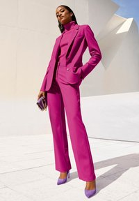Fuchsia blazer and matching wide-leg trousers with a ribbed purple turtleneck; paired with lavender heels and a small purple handbag.