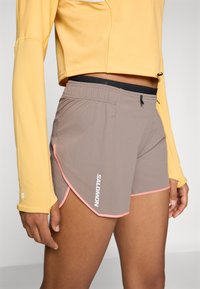 Grå shorts med en texturerad yta, rosa detaljer och svart elastisk midja. Varumärket "SALOMON" tryckt på sidan i vitt.