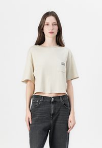 Beige cropped t-shirt met korte mouwen en een borstzak met een grafisch ontwerp. Gecombineerd met hoge zwarte jeans. Effen achtergrond.