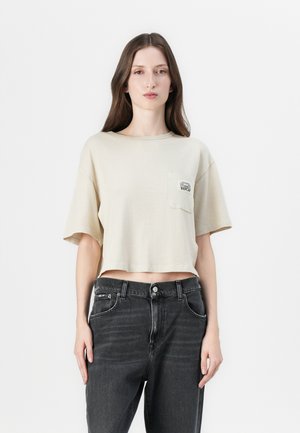 Beige cropped t-shirt met korte mouwen en een borstzak met een grafisch ontwerp. Gecombineerd met hoge zwarte jeans. Effen achtergrond.