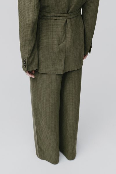 Vue de dos d'une personne portant un costume ample à carreaux vert olive avec une longue veste et un pantalon à jambes larges sur un fond uni.
