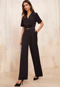 Tuta nera con maniche corte, colletto a rever e cintura con un embellishment circolare. Vestibilità sartoriale e design a gamba larga. Tessuto con texture.