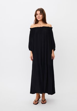 Femme portant une longue robe noire épaules dénudées avec des manches trois-quarts et des sandales noires, debout devant un fond blanc uni.