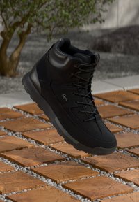 Svart høyre sneakers med lisser på tregulv utendørs, som viser teksturert såle og liten krokodillelogo på siden.