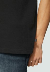 T-shirt noir en tissu texturé, avec une fente sur le côté, et une étiquette de marque bleue visible. Porté avec des jeans en denim bleu.